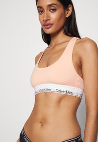 Körüljár önálló barack színű Calvin Klein bralette széles fehér pánttal, fekete logóval; sima anyag, versenypályás hát és minimalista stílus.