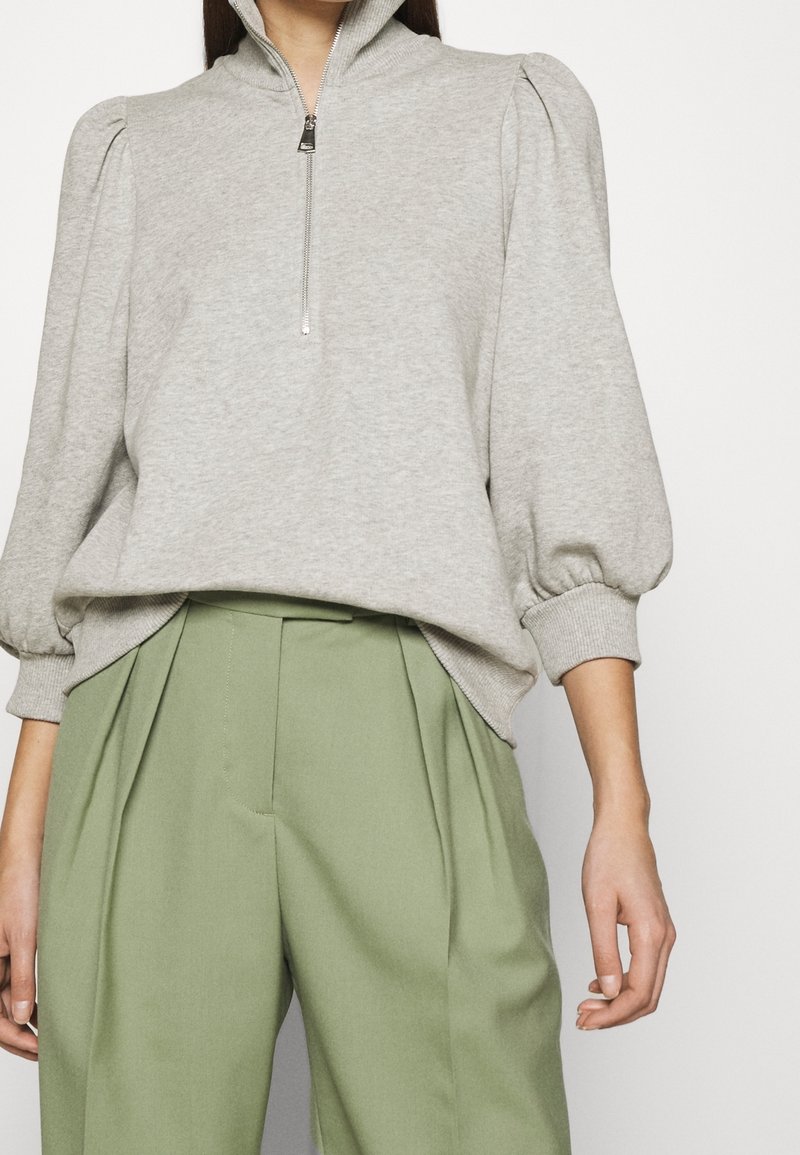 Femme portant un sweat gris clair à fermeture quart de zip avec manches bouffantes et pantalon plissé taille haute vert olive.