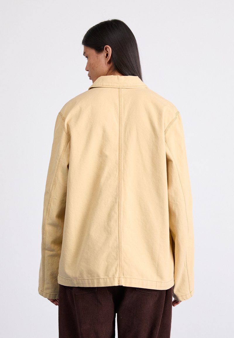 Chaqueta de color amarillo claro, de corte oversized, con un collar sencillo, mangas largas y costuras visibles; combinada con pantalones de pana marrones.