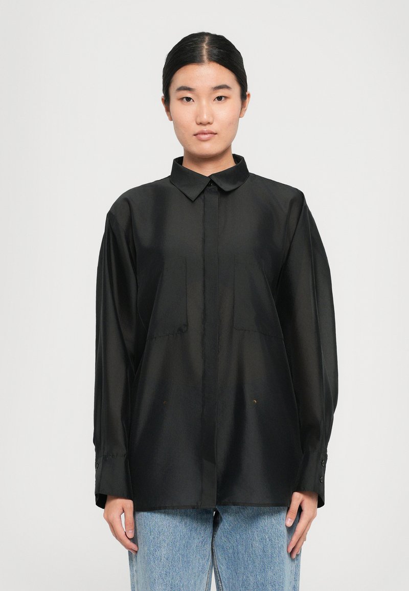 Young woman with straight black hair in a sleek bun wearing a loose black button-up shirt and light blue jeans against a plain background.