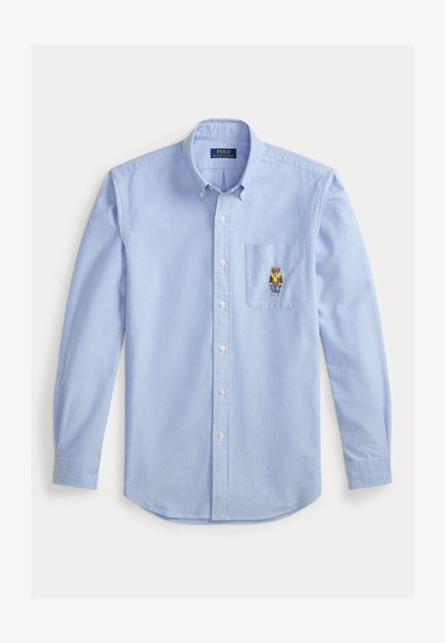 Gai zila krekliņa, kas izgatavots no kokvilnas, dizains ar krūšu kabatu, kurā ir izšūts lāča attēls. Garas piedurknes ar pogu aprocēm.