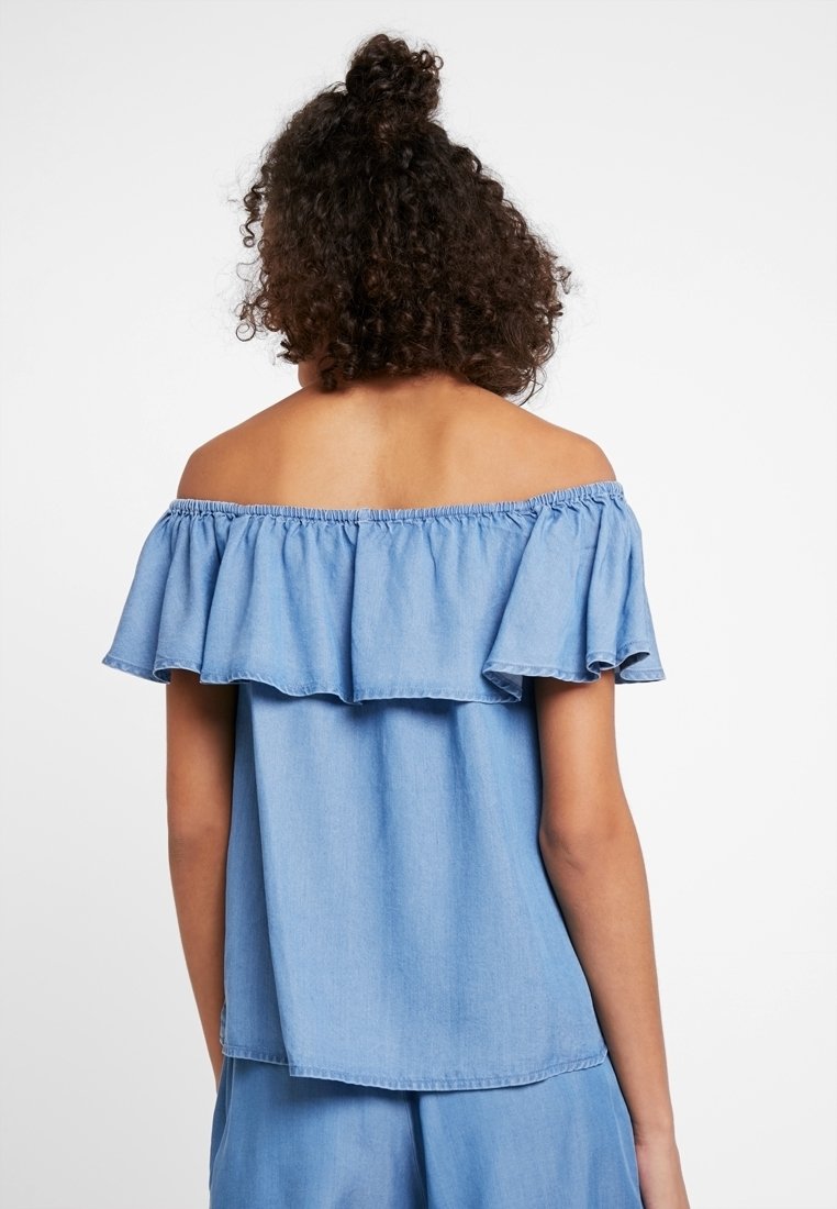 Off-shoulder denimblus i ljusblå, med en volangkrage och lös passform, korta ärmar och en böjd nederkant.
