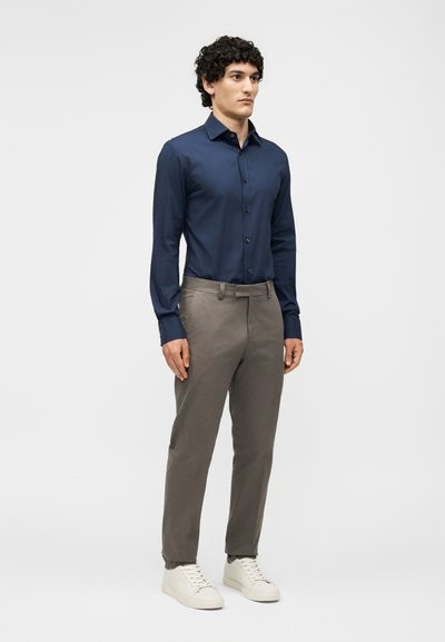 Άνδρας που στέκεται φορώντας ένα μπλε πουκάμισο με κουμπιά, γκρι παντελόνι με θηλιές για ζώνη και λευκά αθλητικά παπούτσια σε ανοιχτό γκρι φόντο.