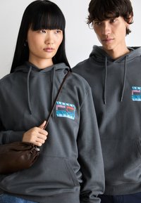 Grijze hoodies met een voorzak en koordjes, voorzien van een multicolore logo op de borst. Eén persoon houdt een bruine tas vast.