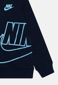 Sudadera de Nike en azul marino, material de algodón, con gran texto "NIKE" en azul claro y gráfico de swoosh, puños y dobladillo acanalados.