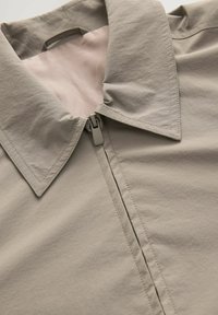 Chaqueta gris claro con cuello puntiagudo, cierre mediante cremallera y textura suave. Detalles de costura claramente visibles y forro en un tono más claro.
