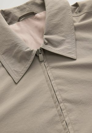 Chaqueta gris claro con cuello puntiagudo, cierre mediante cremallera y textura suave. Detalles de costura claramente visibles y forro en un tono más claro.