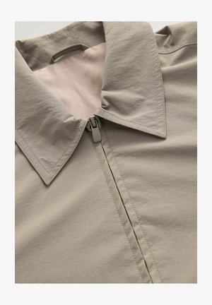 Hellgraue Jacke mit spitzem Kragen, Reißverschluss und glatter Textur. Deutlich sichtbare Nahtdetails und ein Futter in einem helleren Farbton.