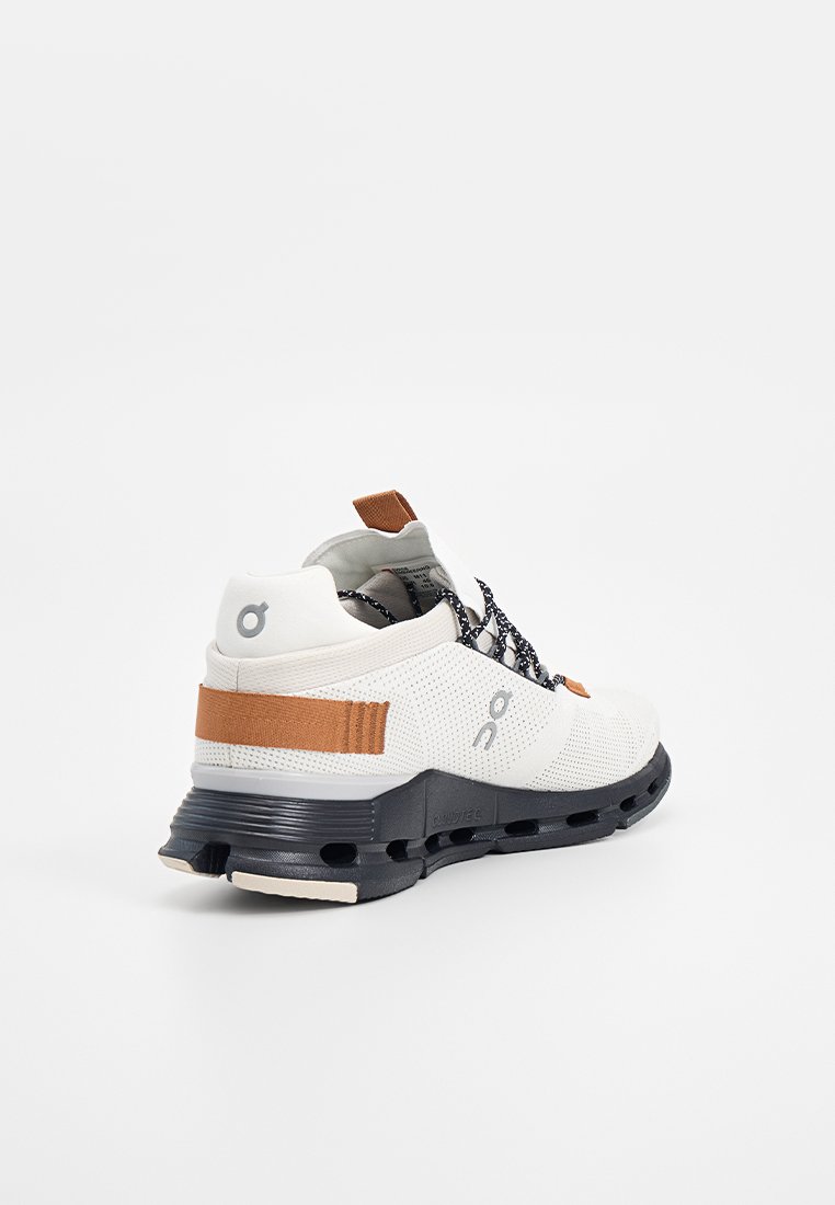 Weißer Sportschuh mit schwarzer Sohle, braunem Fersenriemen und Ziehschlaufe, schwarzem und weißem Schnürsenkel, schräg gezeigt, um die Außenseite und die Ferse zu zeigen.