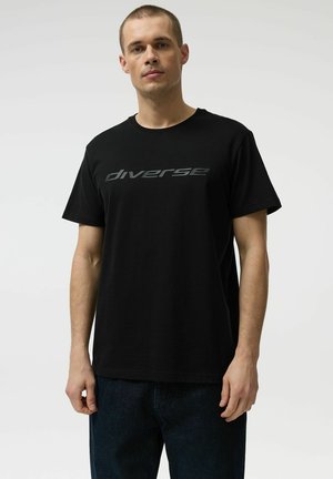 Mężczyzna stojący, ubrany w czarną koszulkę z napisem "diverse" na piersi oraz ciemnoniebieskie dżinsy, na jednolitym jasnym tle.