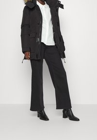 En person står klädd i en svart vadderad jacka med en foder i päls, en vit skjorta och svarta byxor med vida ben, tillsammans med svarta ankelboots.