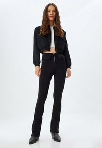 Veste noire coupée avec des boutons-pression et des poches ; associée à un pantalon noir taille haute avec une fermeture éclair à l'avant et des détails de surpiqûres sur les côtés.