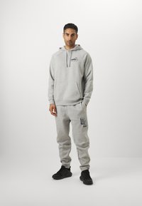 Grå sweatshirt med hætte, kengurulomme og logo-detalje. Sammen med grå joggers med trykt logo. Sorte sportssko.