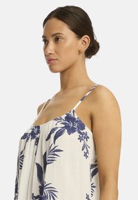 Frau im Profil, die ein blumengemustertes Kleid mit dünnen Trägern trägt, sanfte Züge zeigt und nur minimalen Schmuck trägt.