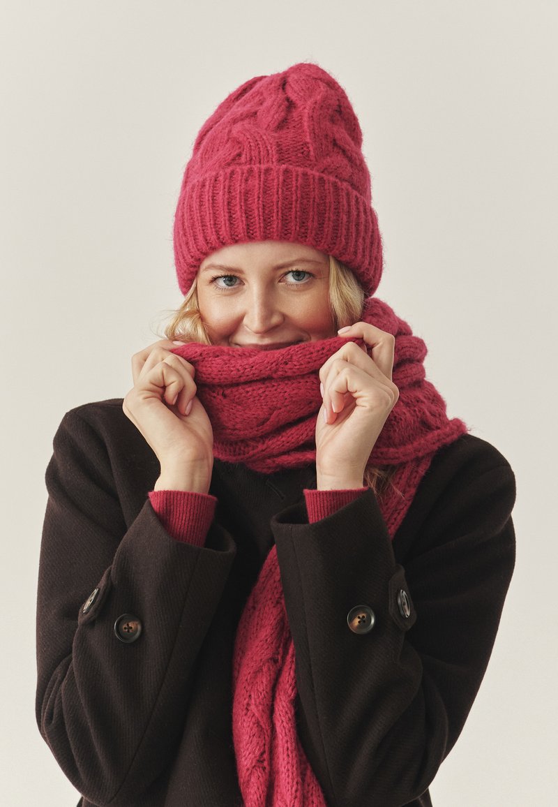 Gebreide roze muts en sjaal set met een geribbelde textuur. Model draagt een zwarte jas met knoopdetails. Gezellige en warme winteraccessoires.