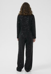 Tuta nera con paillettes, maniche lunghe e gambe larghe, caratterizzata da una superficie testurizzata formata da piccole paillettes per un effetto scintillante.