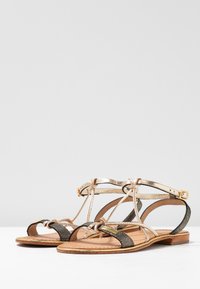 Sandali piatti metallici oro e neri con design a T, realizzati in pelle, con accento annodato e fibbia regolabile alla caviglia.