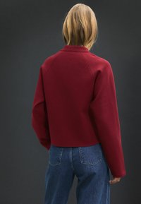 Sweatshirt bordeaux raccourci avec un col haut et des manches raglan, associé à un jean en denim bleu. Texture lisse, détails minimalistes.