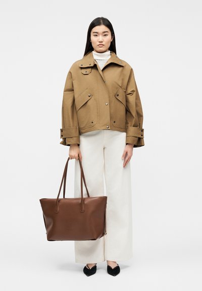 Γυναίκα που φοράει καφέ μπεζ μπουφάν, λευκό παντελόνι με φαρδιά πόδια, μαύρα μυτερά παπούτσια και κρατά μια μεγάλη καφέ δερμάτινη τσάντα ώμου.