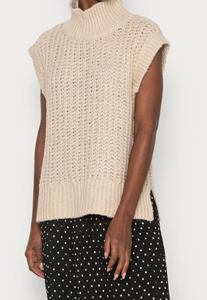 Pull sans manches en maille beige à col roulé, porté sur une jupe noire à pois blancs, laissant apparaître les bras et le torse.