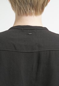 Czarna koszula z prostym dekoltem, widocznymi szwami na ramionach oraz małą metką oznaczoną „R.A.W.” z tyłu.