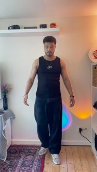 Top preto sem mangas, jeans escuros largos e ténis cinzentos. Colar e pulseiras prateados em destaque. Chão de madeira com luzes de parede coloridas.