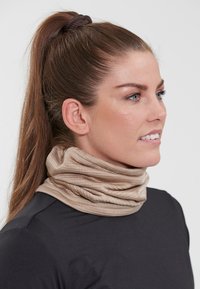 Beige stickad halsduk med en texturerad, åtsittande design, hängande runt halsen. Bärs över en svart långärmad skjorta.