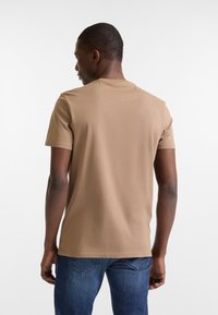 Camiseta marrón de manga corta con cuello redondo clásico, hecha de suave algodón; modelo mostrado de espaldas, combinado con jeans azules.