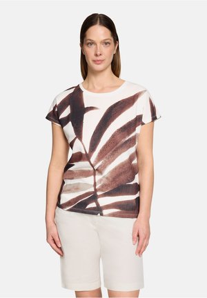 Frau mit glatten braunen Haaren, die ein weißes kurzärmliges Oberteil mit großem braunem Blattmuster und weiße knielange Shorts trägt.