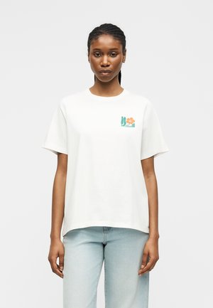 Vrouw met gevlochten haar draagt een wit T-shirt met korte mouwen met een klein oranje bloem- en groen bladmotief op de linkerborst en lichtblauwe spijkerbroek.