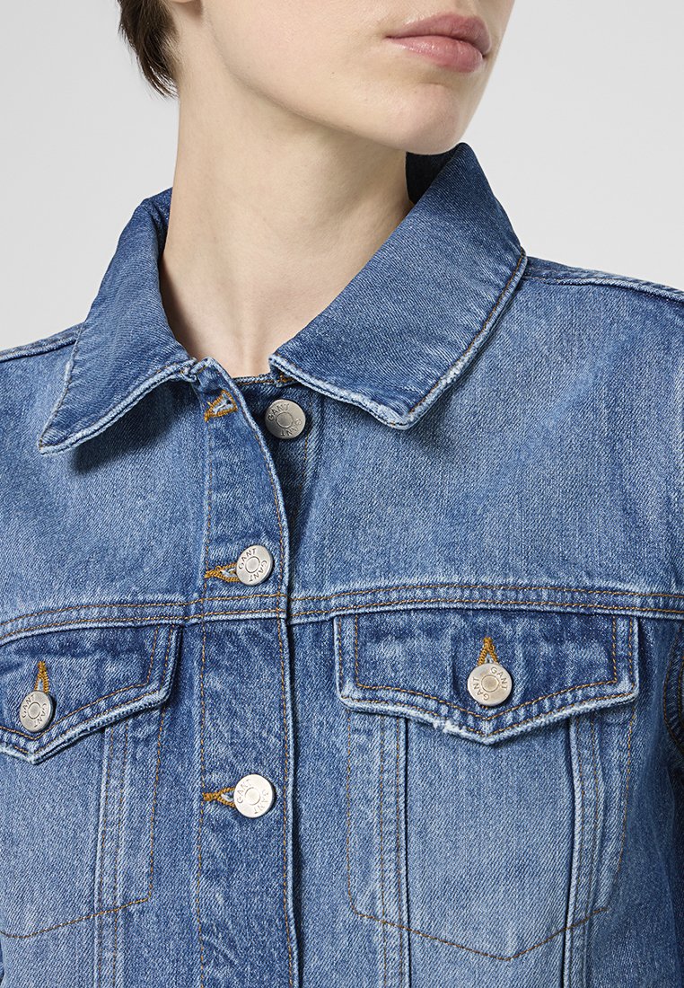 Jeanjacke in mittlerem Blau mit einem klassischen Kragen, silbernen Knöpfen, Brusttaschen und sichtbaren Steppnähten. Der Stoff hat eine weiche Textur.