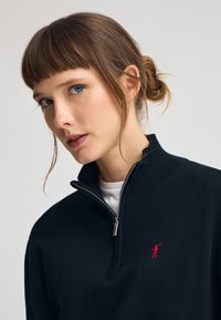 Marinblå tröja med halvdragkedja i stickad textur och silverfärgad dragkedja. Anmärkningsvärt röd broderad logotyp på vänster bröst. Jerseylager under.