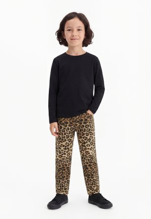 Enfant aux cheveux mi-longs et foncés, portant un t-shirt noir à manches longues, un pantalon imprimé léopard et des baskets noires, debout avec une main dans la poche.