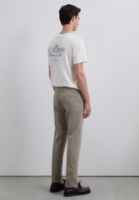 Pantalones de algodón beige y una camiseta crema con texto oscuro en la espalda. El modelo está de lado, mostrando claramente el ajuste y los detalles del diseño.