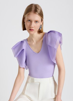 Giovane donna con capelli biondi corti che indossa una maglietta viola chiaro con maniche arricciate e pantaloni bianchi a vita alta su uno sfondo semplice.