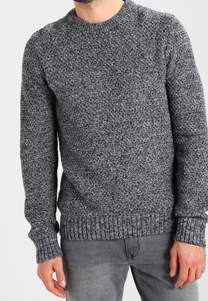 Mann trägt einen strukturierten dunkelgrauen Strickpullover mit gerippten Bündchen, Saum und Ausschnitt, kombiniert mit mittelgrauen Jeans.
