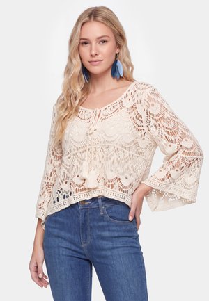 Ung kvinde med langt blondt hår iført en cremefarvet blondebluse med klokkeærmer og blå jeans, poserer med hånden på hoften.