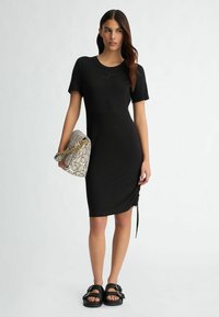 Black short-sleeve fitted dress with a rounded neckline, featuring gathered side detailing. Paired with a patterned handbag and chunky sandals.