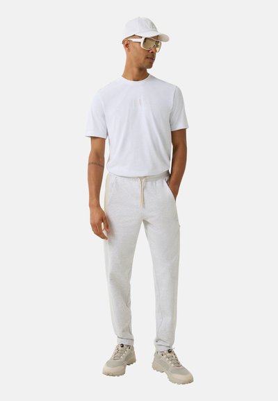 Weißes T-Shirt mit kurzen Ärmeln, graue Sweatpants mit elastischem Bund und beigen Sneakers. Die Accessoires umfassen eine weiße Kappe und übergroße weiße Sonnenbrillen.