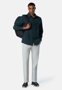 Camisa abotonada de color verde oscuro con dos bolsillos en el pecho, pantalones claros grises, mochila negra y zapatos oscuros con suela texturizada.