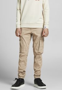 Beige cargo kalhoty se zúženými manžetami, několika kapsami a měkkou bavlněnou směsí. Nesené s svetrem ve světlé barvě a černými teniskami.