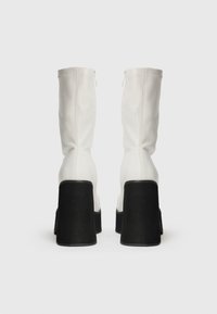 Boots albe până la genunchi, cu un design ajustat, tocuri groase negre, textură netedă și închidere cu fermoar vizibil la spate.