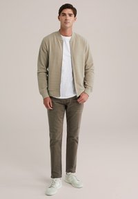 Chaqueta bomber texturizada color beige claro sobre una camiseta blanca, combinada con pantalones marrones y zapatillas blancas. Diseño simple y moderno.