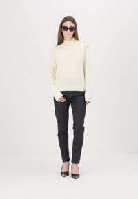 Maglione a coste crema con collo alto e dettaglio di bottoni dorati su una spalla, abbinato a jeans neri aderenti e tacchi a punta neri.