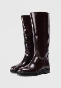 Γυαλιστερά μπορντό μποτάκια μέχρι το γόνατο με στρογγυλή μύτη, επίπεδη μαύρη σόλα και αντίθετες κόκκινες ραφές κατά μήκος των ραφών.