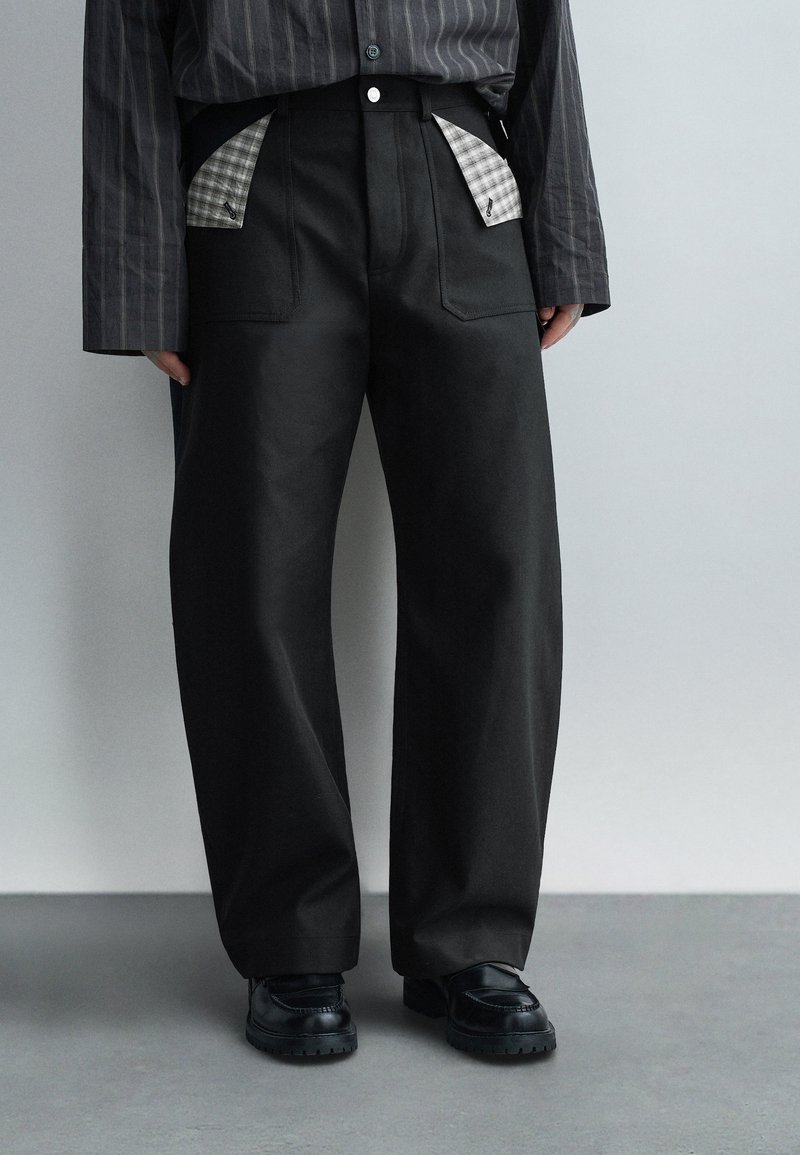 Osoba ubrana w czarne spodnie o szerokich nogawkach odsłaniające wstawki w kratkę przy kieszeniach, w połączeniu z czarnymi mokasynami i ciemną koszulą w cienkie prążki.