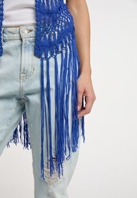 Blå makraméväst med långa fransdetaljer, bärs över en vit topp och ljus tvättade slitna jeans, med en avslappnad passform och öppen väv.