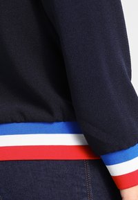 Marineblaues Stoffjackett mit geripptem Saum, der rot, weiß und blau gestreift ist. Die strukturierte Oberfläche und elastische Bündchen sind sichtbar.
