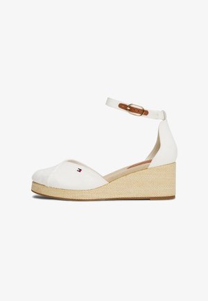 Zapato cuña blanco de lona con tira en el tobillo, hebilla marrón, tacón de textura trenzada y pequeño logo en rojo, blanco y azul en la parte superior.