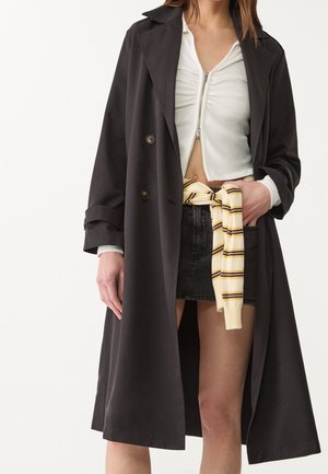 Vrouw draagt een lange zwarte jas over een witte cropped top met rits, een zwarte spijkerrok en een geel gestreepte trui om haar middel geknoopt.